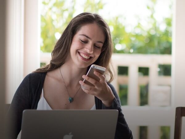 A woman smiling at her iphone and sitting in front of her laptop
